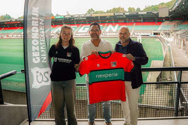 Een medewerker van GezondRijk Nijmegen en twee medewerkers van Kompas Veiligheidsgroep houden samen een rood-groen N.E.C.-shirt omhoog op de tribune van het stadion van N.E.C., naast een banner van GezondRijk Nijmegen.